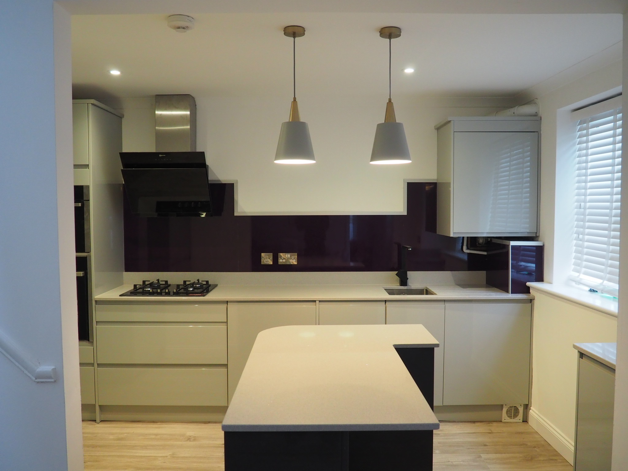 Modern kitchen featuring sleek grey cabinetry, a gas stovetop under a stainless steel range hood, and a black sink with a purple backsplash. An island with a light countertop sits centrally, illuminated by two pendant lights. Supported by expert building services, a window with white blinds is on the right.