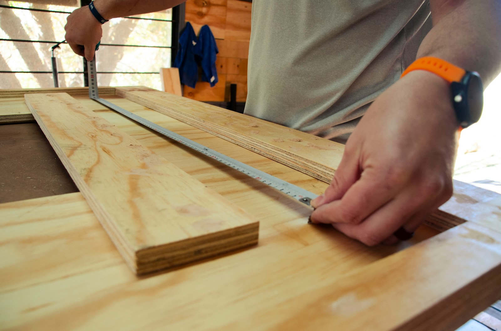 Close-up of a person using a metal ruler and pencil to measure and mark a piece of wood on a workbench for building services. The person is wearing a smartwatch and a gray shirt. The workspace is well-lit with natural light, showcasing a window and a blue jacket in the background.
