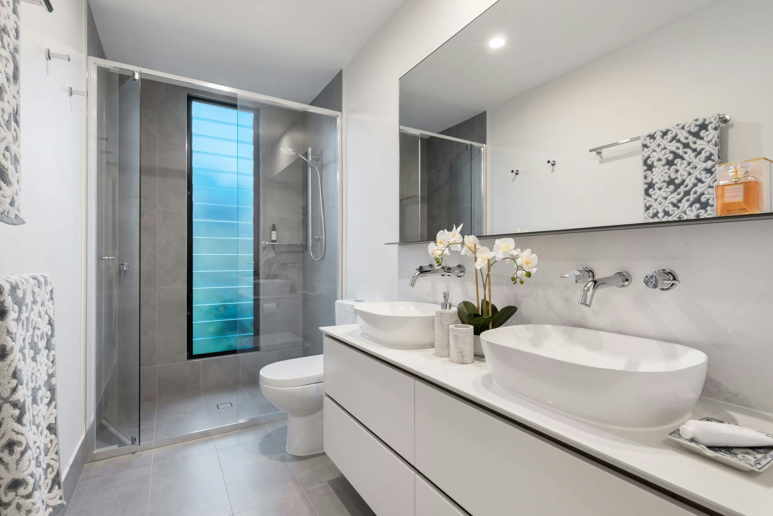 A modern bathroom with a glass-enclosed shower, a white toilet, a large wall mirror, and a dual sink vanity with round white basins. The countertop features a small potted orchid and a soap dispenser. Building services have ensured the room has a clean, minimalist design with gray accents.
