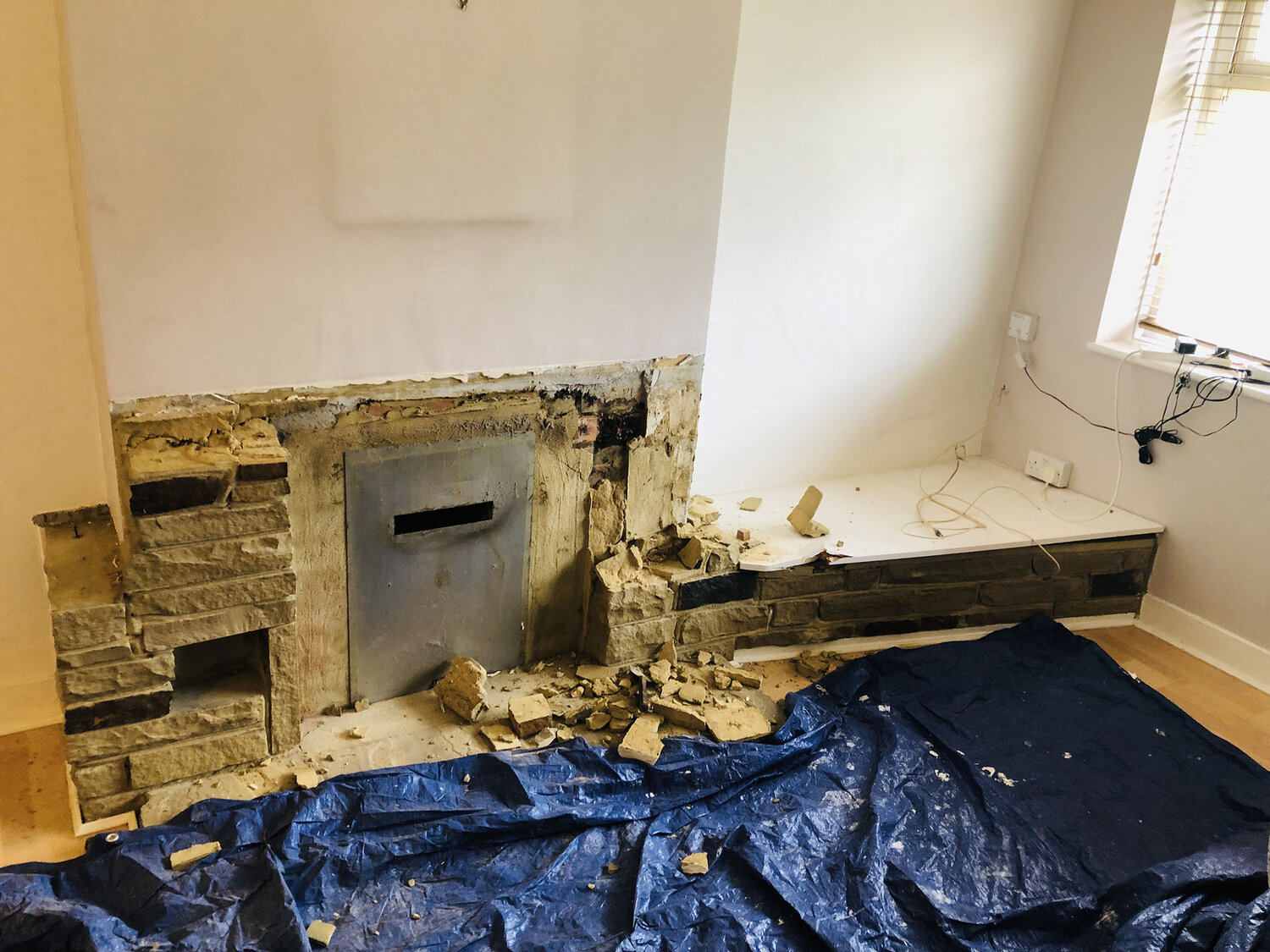 A partially demolished fireplace with exposed bricks and rubble scattered on the floor. A blue tarp covers part of the wooden floor. Nearby, a white wall and a window with blinds are visible. Cables are hanging by the wall.