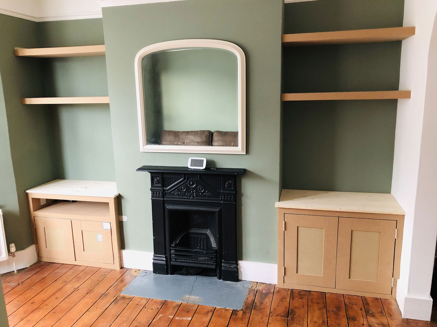 A living room with green walls, wooden floor, a black fireplace, a mirror above it, and built-in wooden cabinets with open shelves on either side of the fireplace.