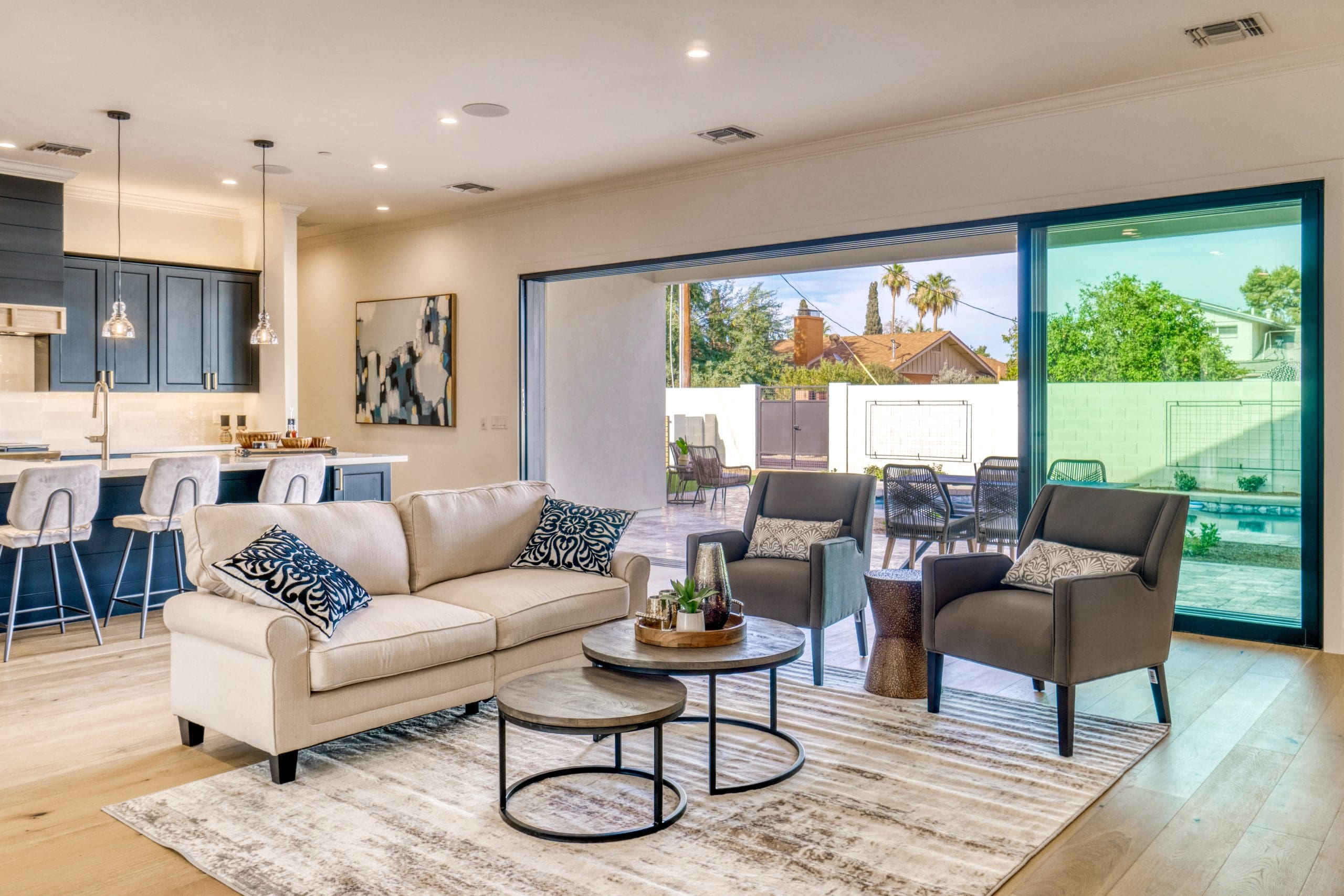 A modern open-concept living room with a beige sofa, two gray armchairs, nested coffee tables, and a rug. Large sliding doors open to a patio with outdoor seating and greenery. Kitchen with blue cabinets is visible in the background.