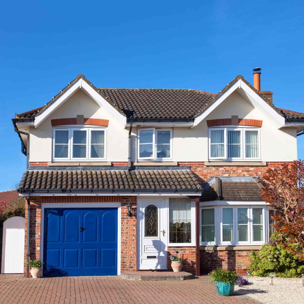 A two-story brick and cream house with a dark tiled roof, blue garage door, white front door, and large windows. There are potted plants and a tree in the front yard under a clear blue sky.