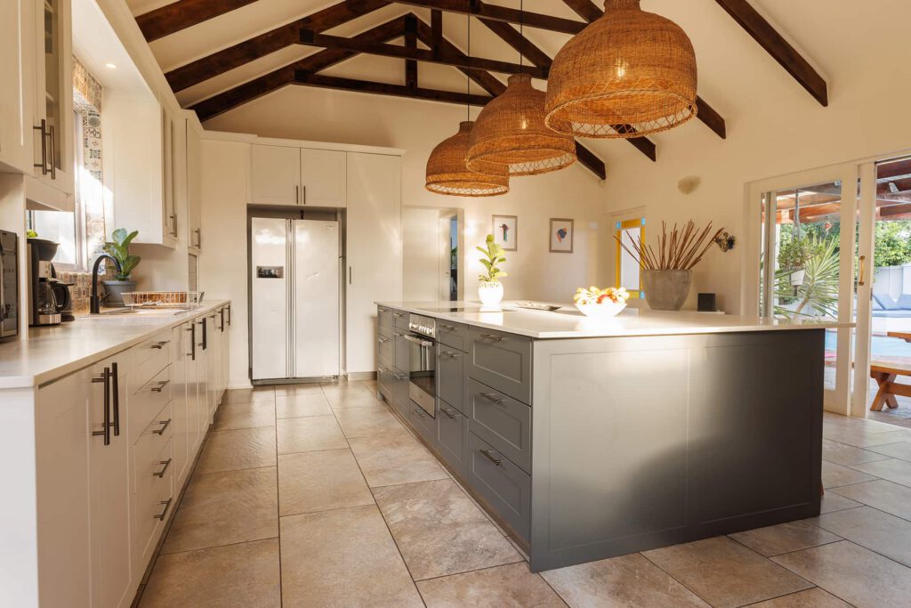 Modern kitchen with exposed wooden beams, large island with gray cabinets, wicker pendant lights, stainless steel appliances, and sunlit tile flooring. Plants and decorative items add warmth to the bright, airy space.