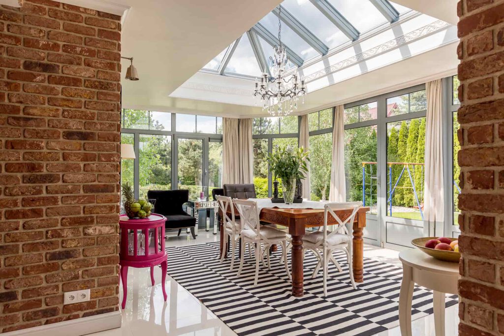 A sunlit dining room features a glass ceiling, brick walls, striped rug, wooden table with white chairs, a chandelier, and large windows overlooking a green garden.