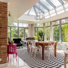 A sunlit dining room features a glass ceiling, brick walls, striped rug, wooden table with white chairs, a chandelier, and large windows overlooking a green garden.