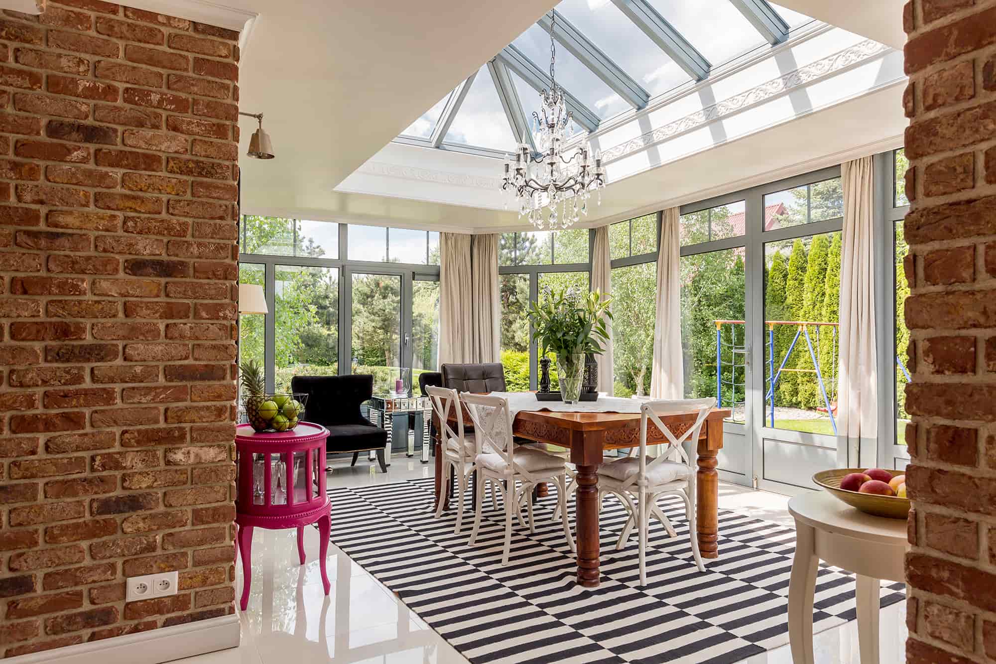 A sunlit dining room features a glass ceiling, brick walls, striped rug, wooden table with white chairs, a chandelier, and large windows overlooking a green garden.